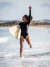 Woman on beach with surfboard in Black Long Sleeve Surf Swimsuit | The Sulawesi Midnight 